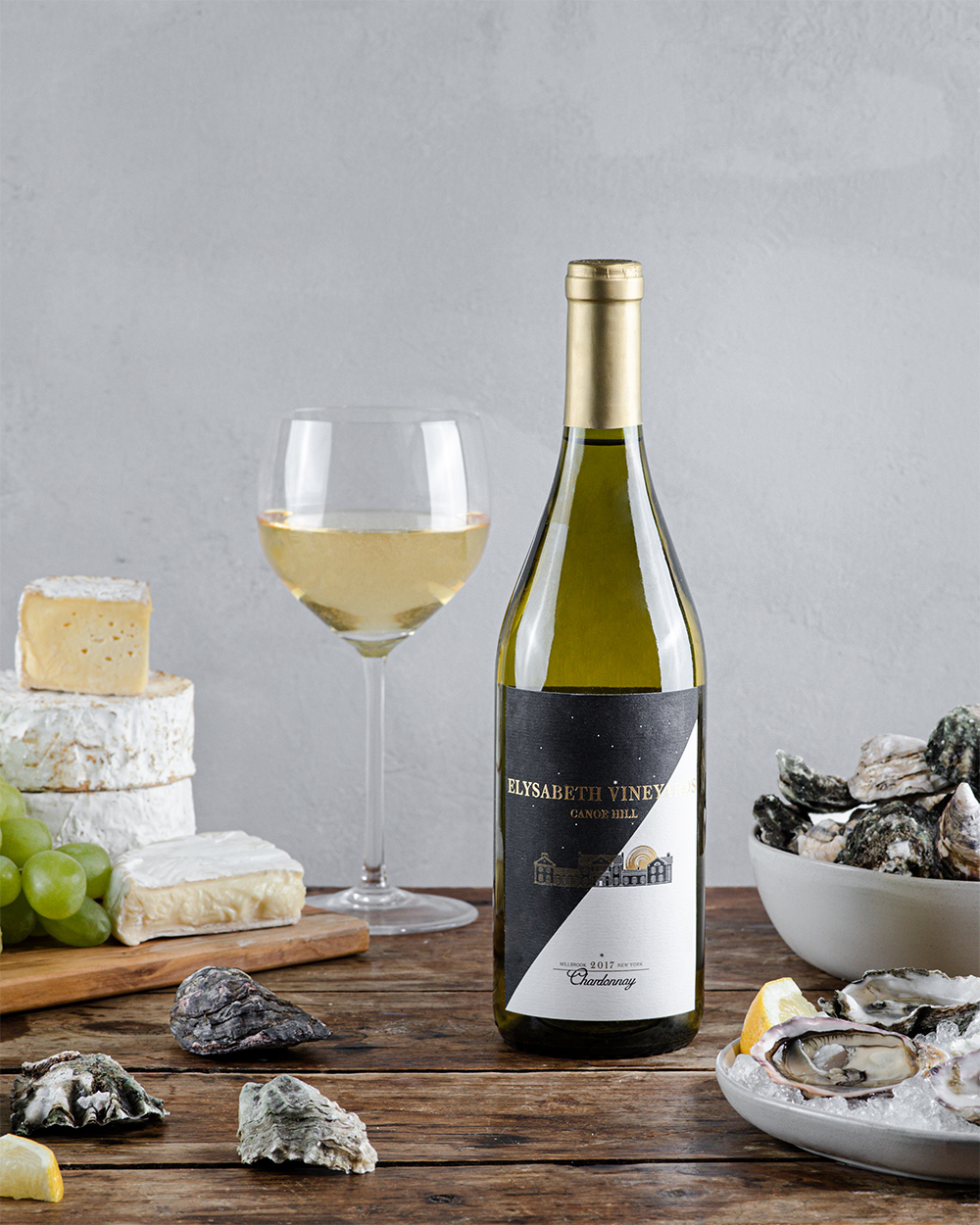 Chardonnay bottle with half full wine glass, surrounded by brie cheese, clams, oysters, and green grapes.